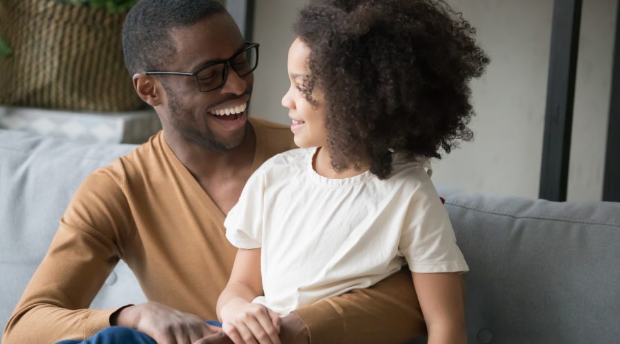 Smiling african American young dad sit on couch hug cute little daughter chatting or talking, happy black father spend time having conversation with funny preschooler child, enjoy weekend together